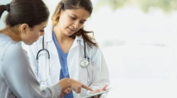 An attentive doctor listens as a patient discusses medical test results. The patient is pointing at something on the doctor's digital tablet.